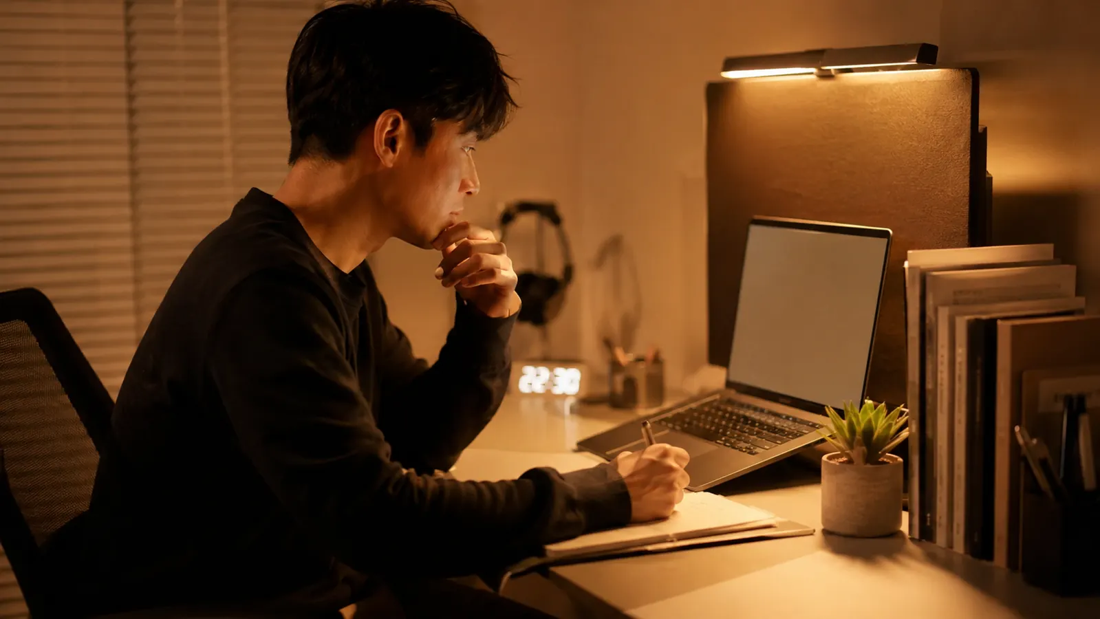 Warm amber light from a monitor light bar illuminating a tidy home office desk at night, with a laptop and notebook visible